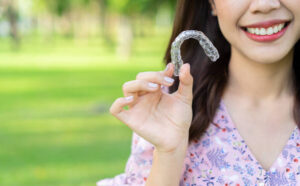 close up young woman smiling hand holding dental aligner retainer (invisible) at outdoor nature park and garden background for beautiful teeth and dental treatment course concept